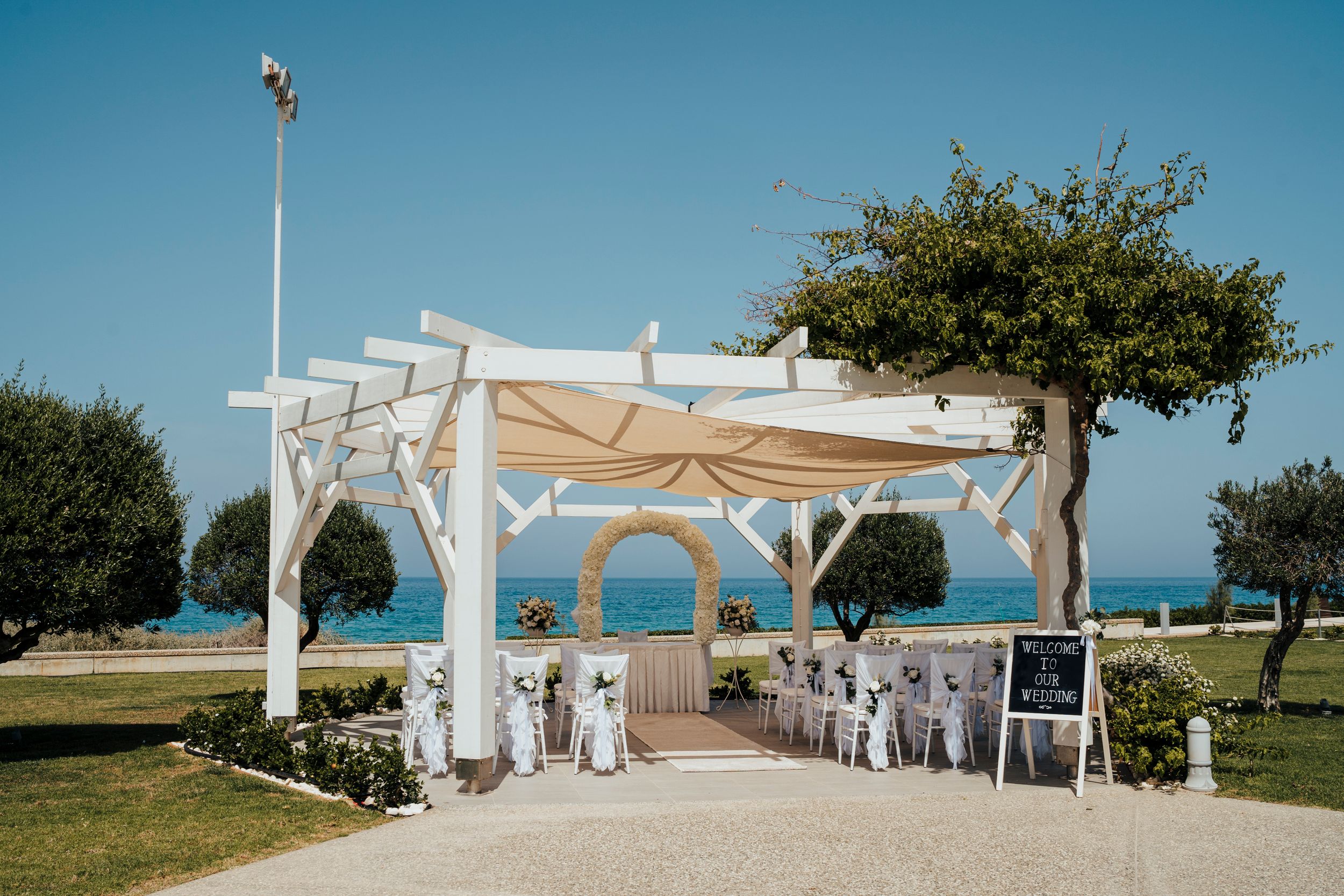 A white wooden wedding pergola decorated with fabric and chairs set up overlooking the ocean on a sunny day.