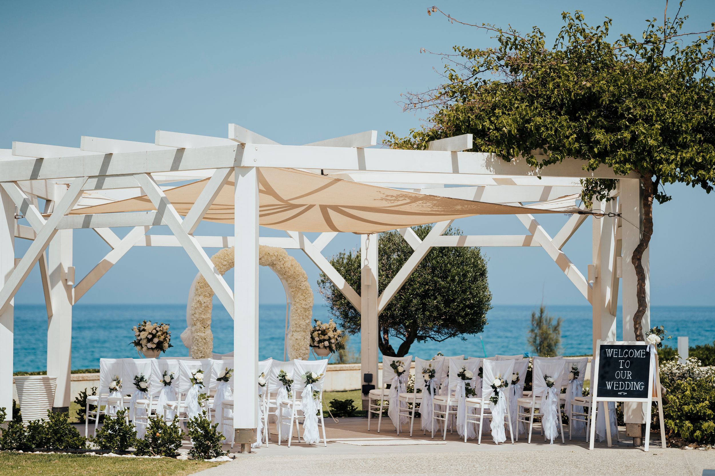 A romantic beach wedding setup with white pergola, draped fabric, and ocean views in the background.