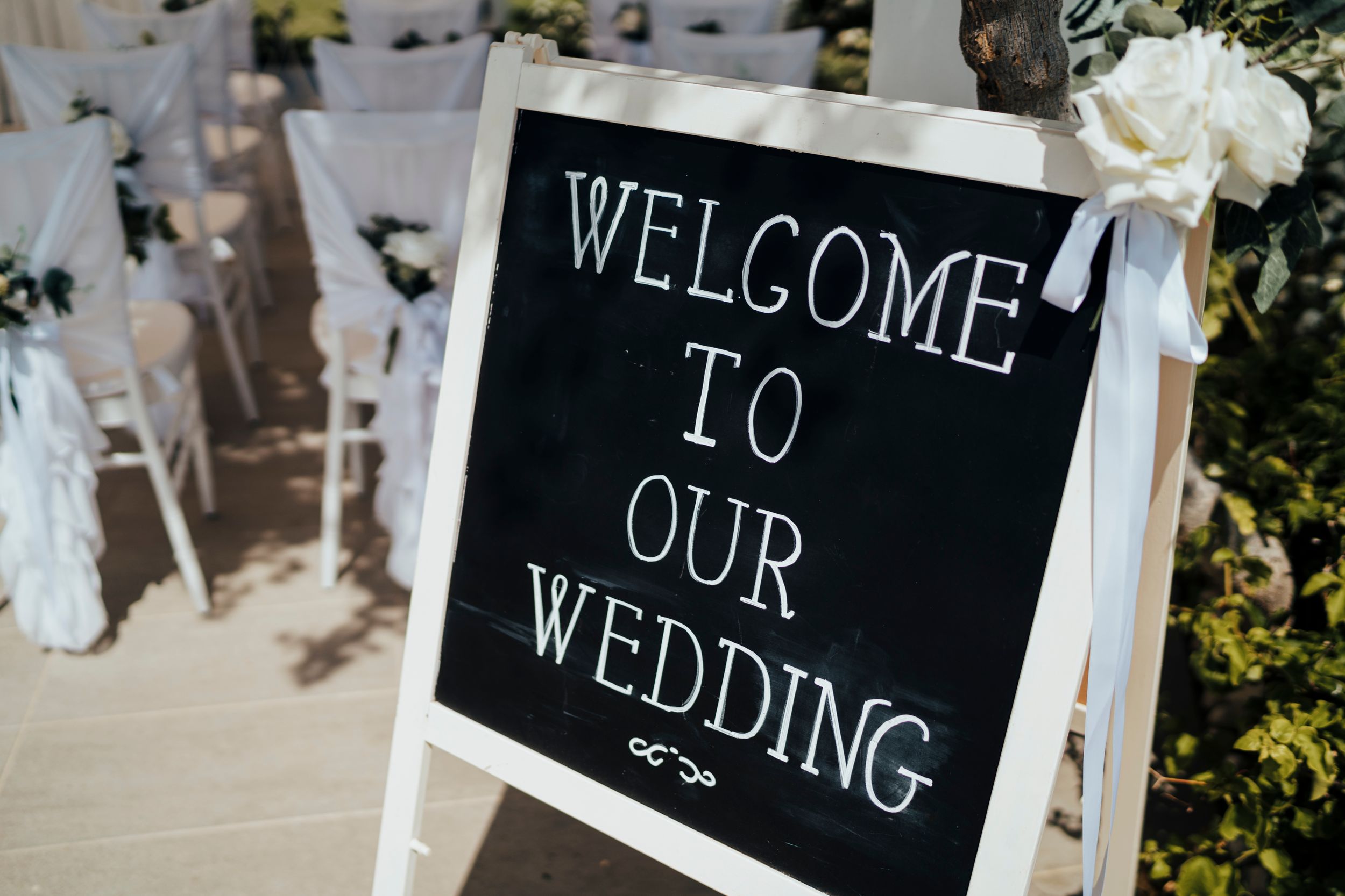 A black chalkboard welcome sign at a wedding ceremony with white chairs and floral decorations in the background.