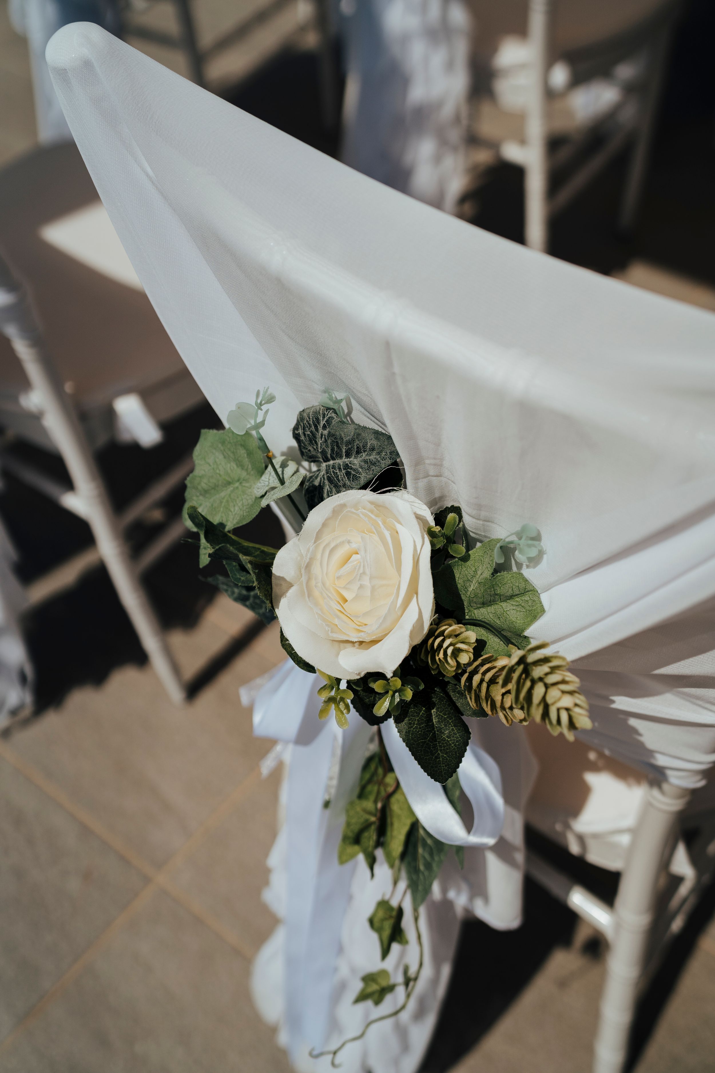 A white rose and greenery arrangement decorates a white fabric chair sash at a wedding ceremony.