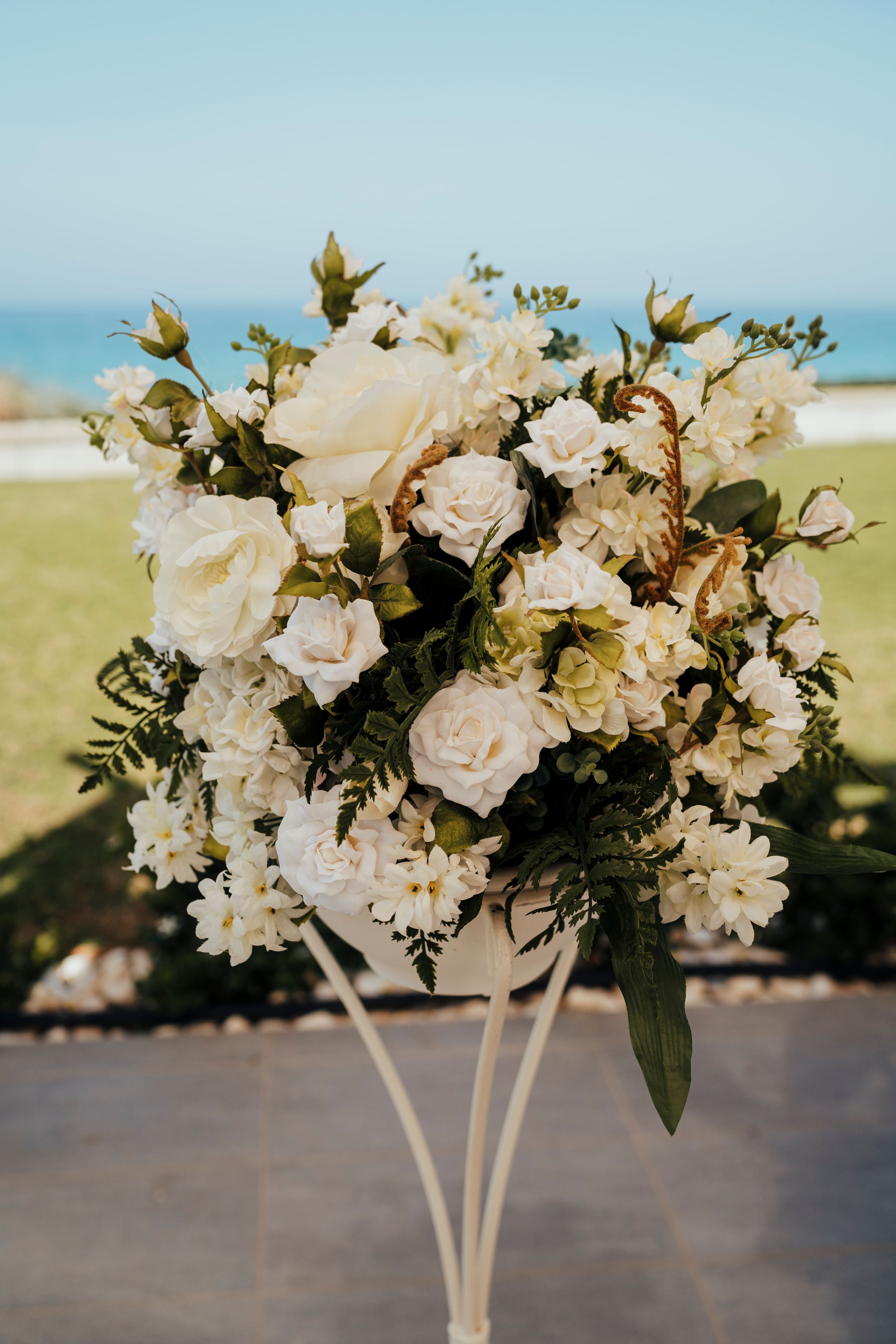 Elegant white floral arrangement with roses and greenery on a stand against a blurry ocean backdrop.