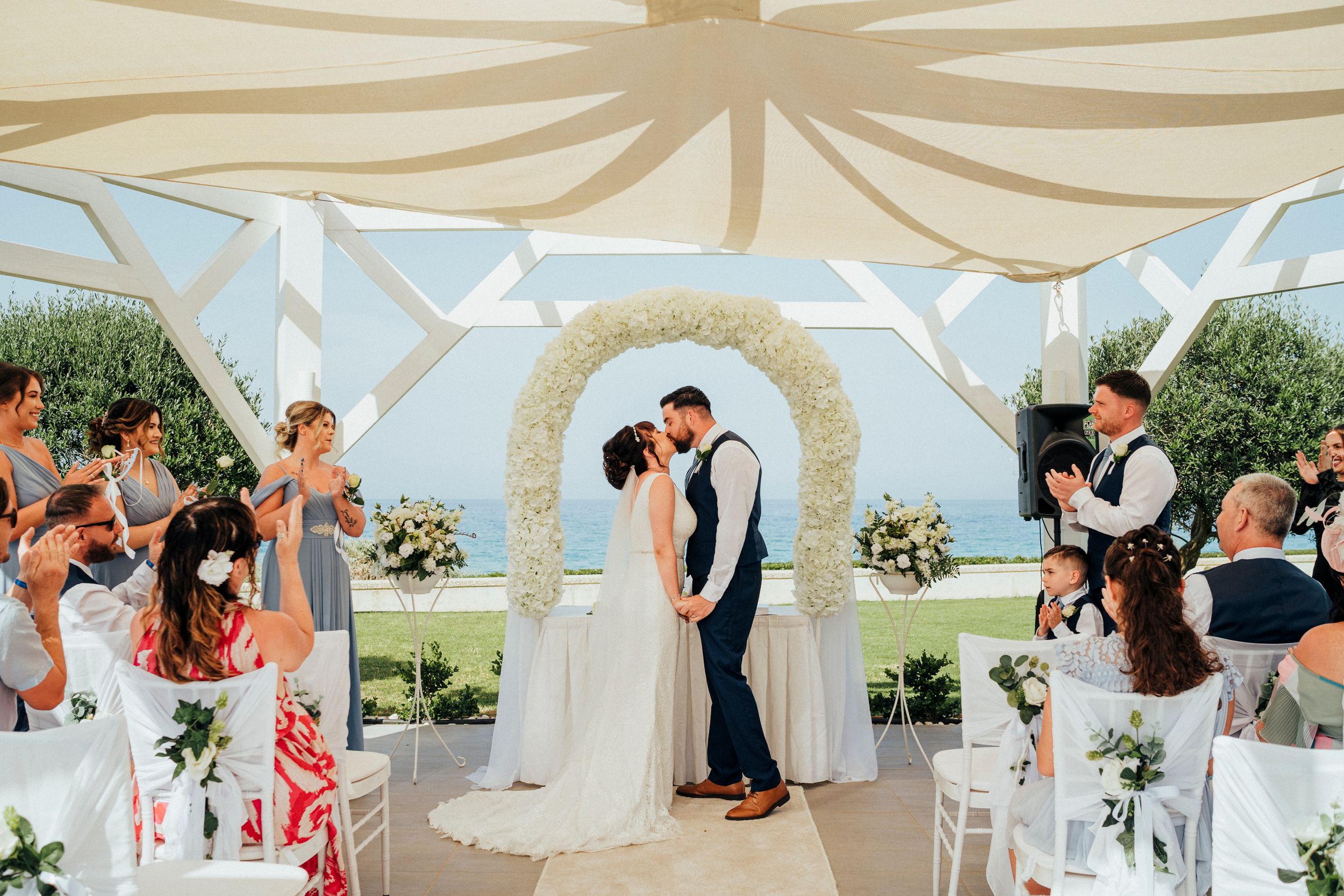 A beautiful outdoor wedding ceremony takes place under a draped gazebo with floral arrangements and gathered guests.