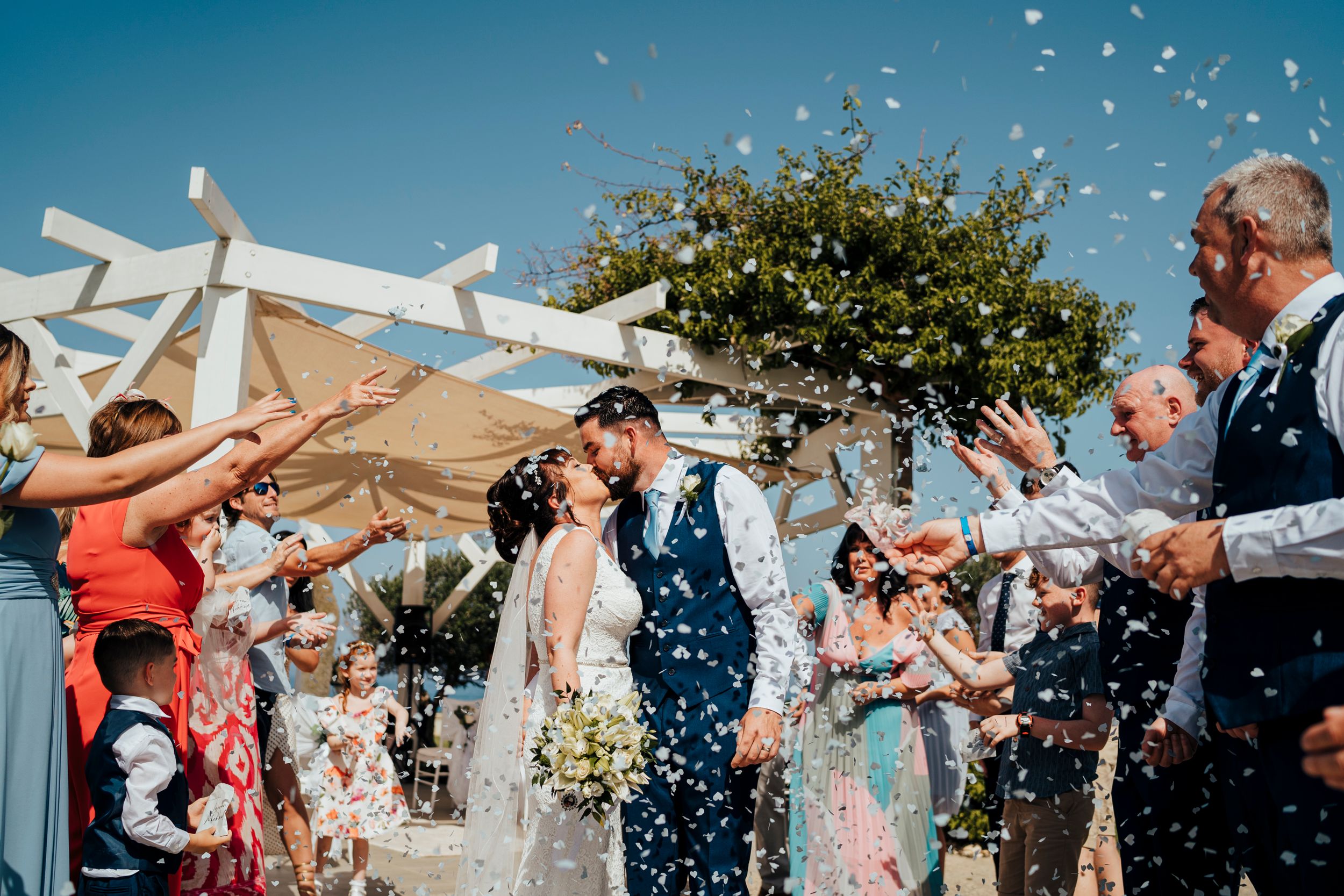 Wedding guests shower a couple with confetti during a sun-filled outdoor ceremony under a white arch.