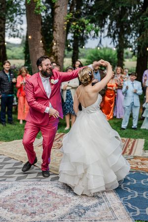A couple dances joyfully at their outdoor wedding celebration on vintage carpets under trees while guests watch and smile.