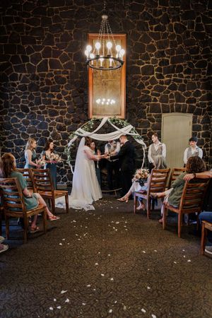 An intimate indoor wedding ceremony in a rustic stone-walled venue with elegant chandeliers and scattered flower petals.