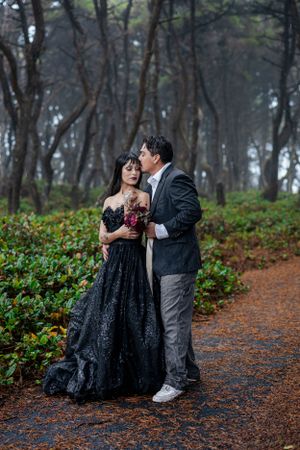 A couple shares an intimate moment on a forest path while one wears a flowing black gown.