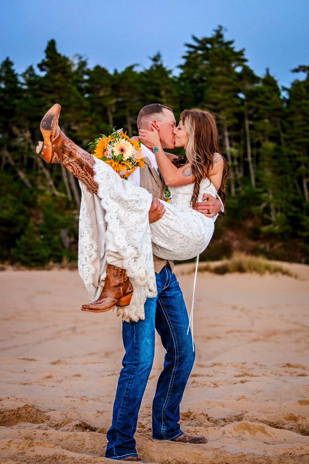 A couple shares a romantic kiss on the beach with evergreen trees in background as she wears cowboy boots and white dress.