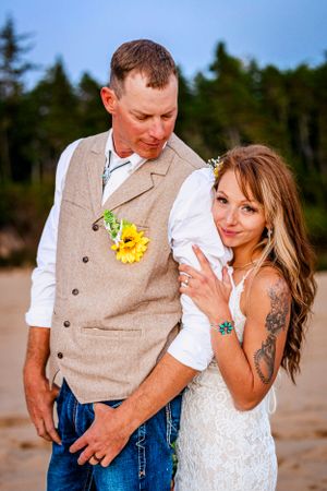 A romantic moment between newlyweds on beach with pine forest backdrop, wearing casual country wedding attire.