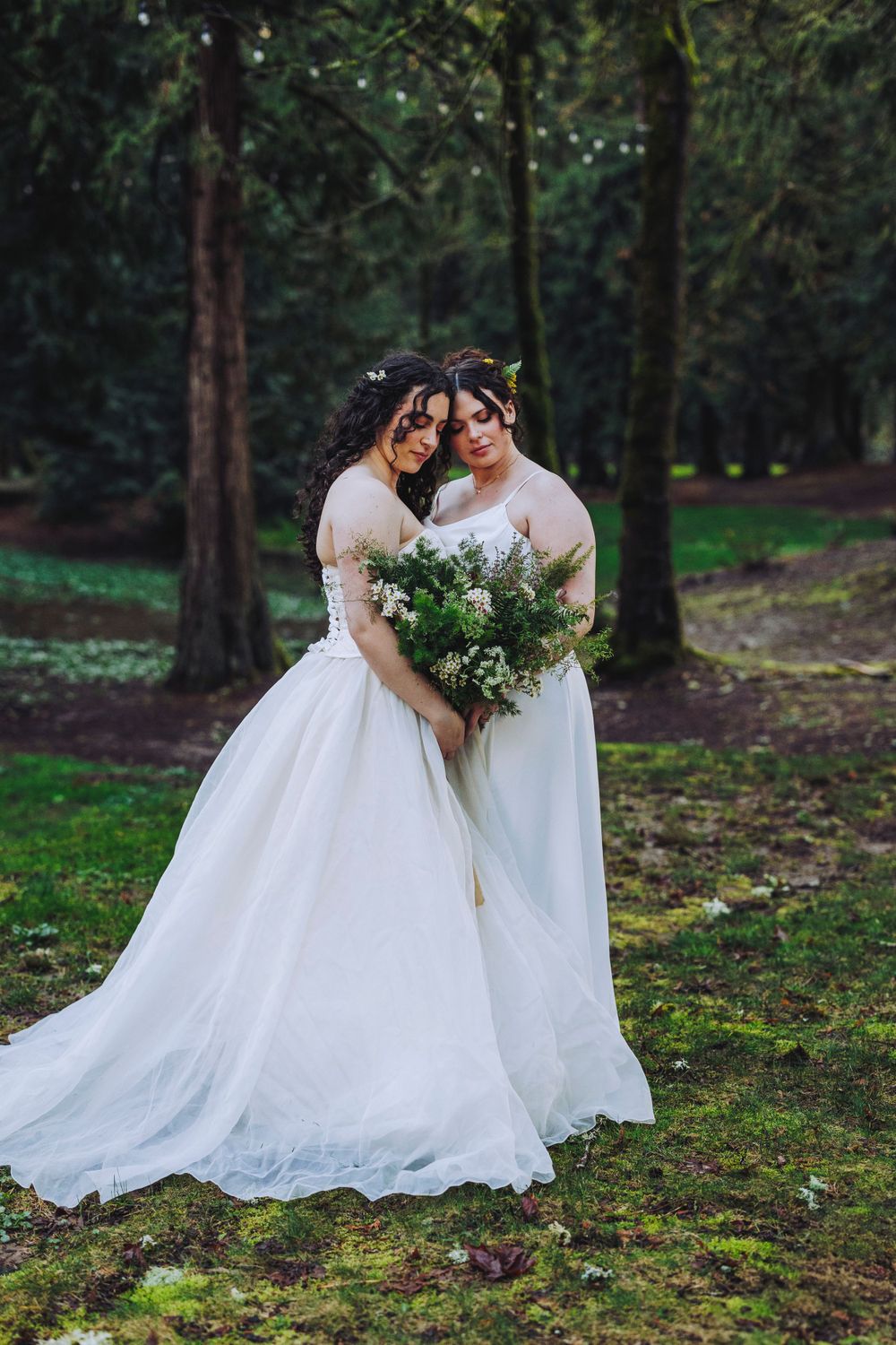 Two brides share an intimate moment in a forest setting while holding lush green and white bouquets.