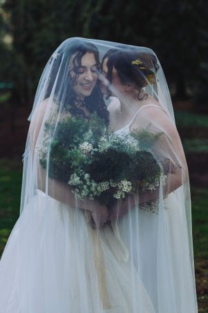 Two figures share an intimate moment under a flowing white veil while holding white floral bouquets in an outdoor setting.