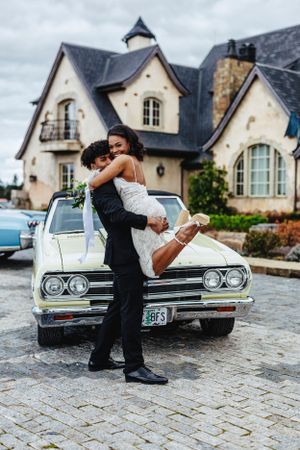 Newlywed couple embracing in front of vintage white car and elegant stone mansion on cobblestone driveway.