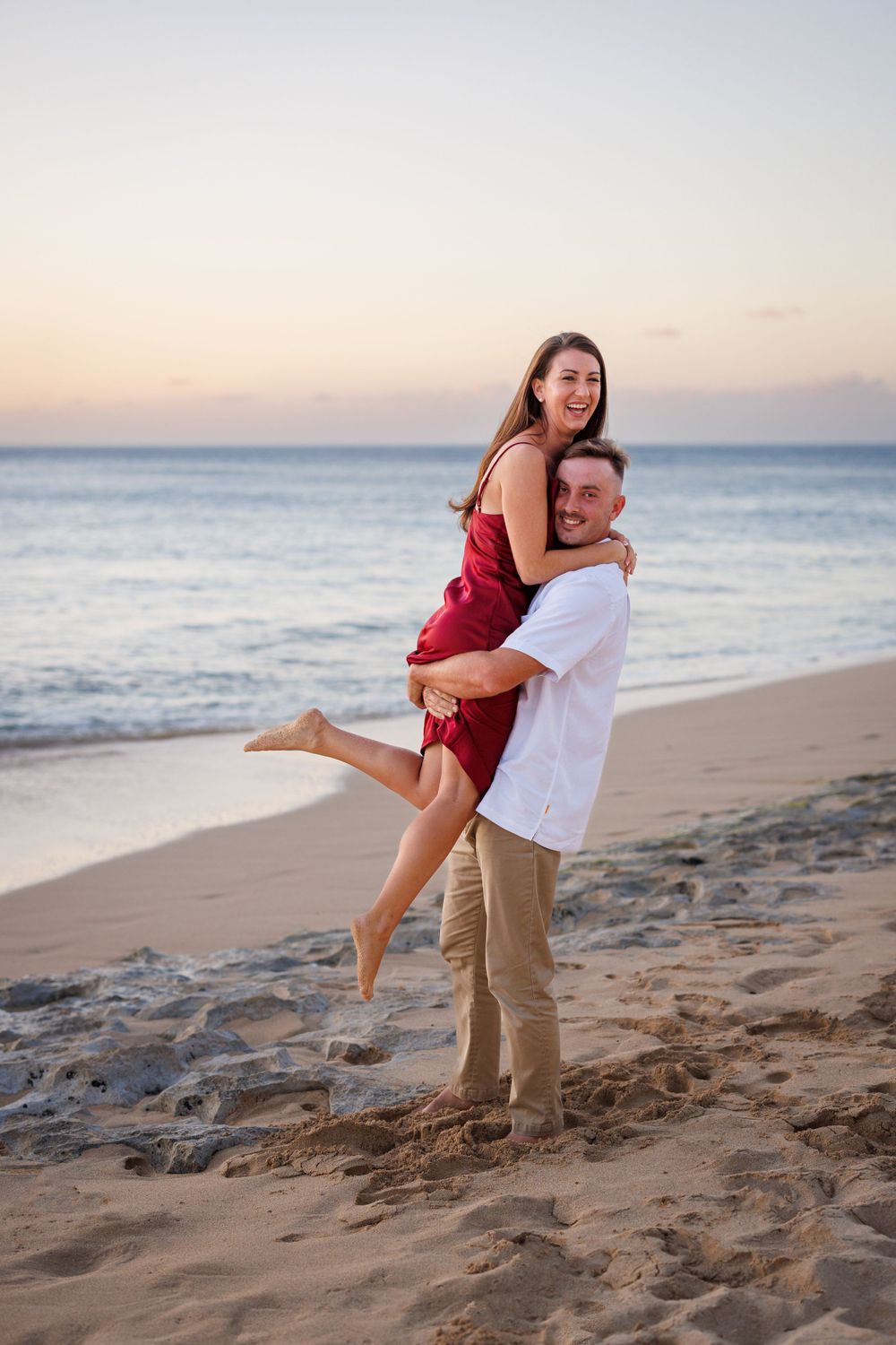 Couple sharing a joyful piggyback ride on a sandy beach at sunset, woman in red dress laughing.