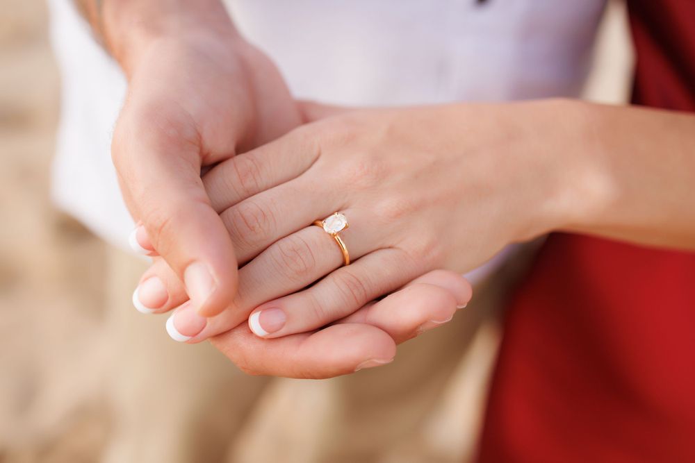 Close-up of a woman's hand wearing a delicate gold engagement ring with a small diamond, held gently by another hand.