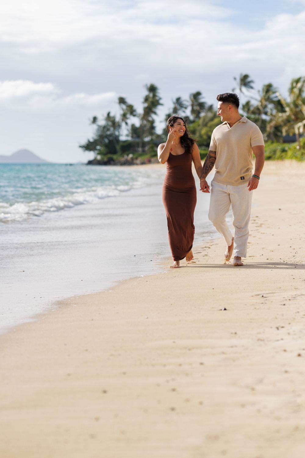 A couple walks hand-in-hand along a tropical sandy beach, with palm trees and ocean waves in the background.