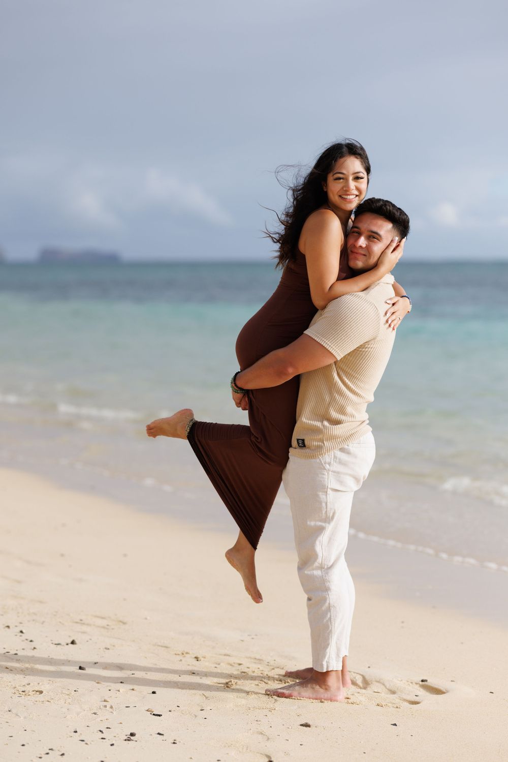 Happy couple embracing on a tropical beach, man lifting woman in brown dress, white sand and turquoise ocean backdrop.
