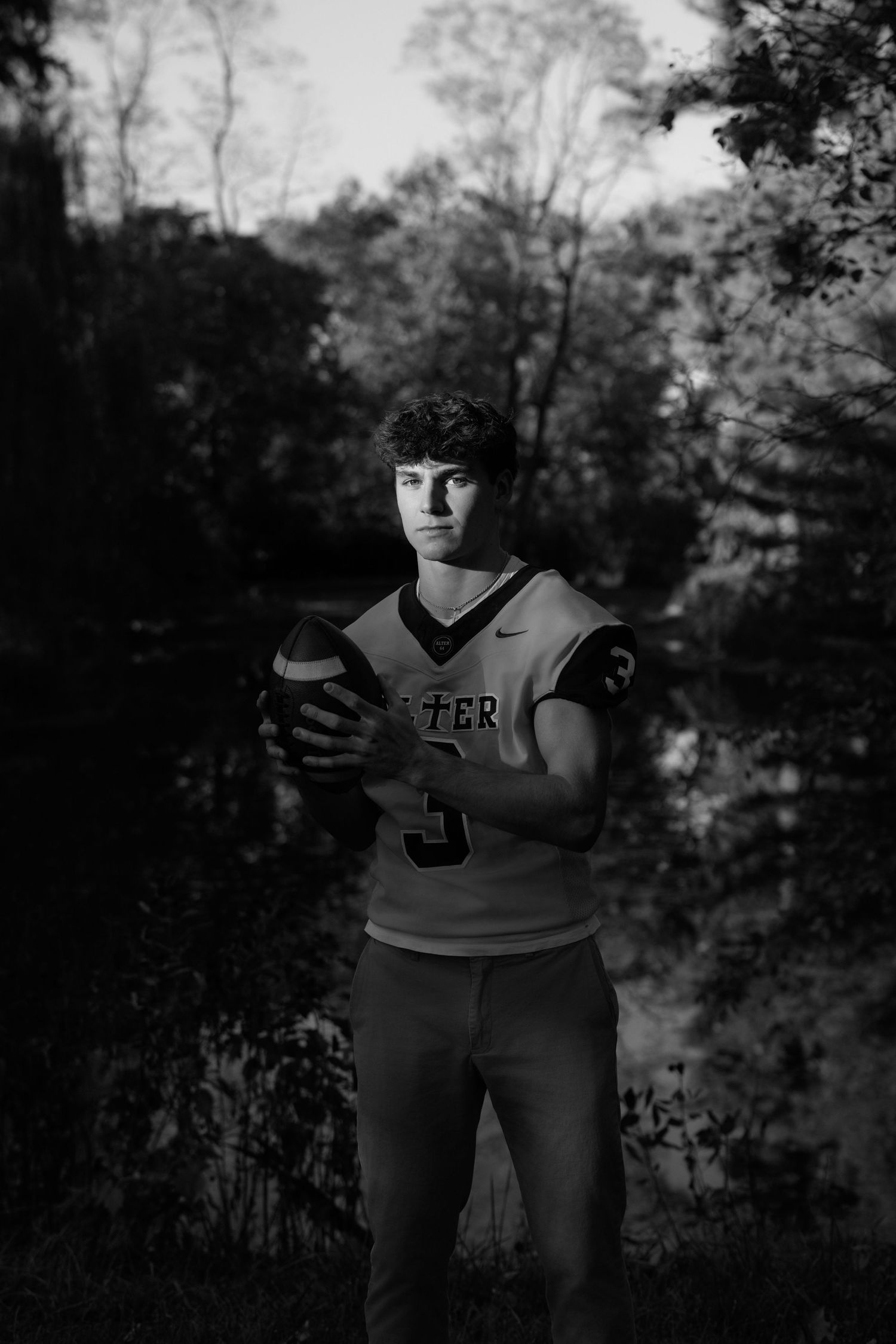 Black and white portrait of a high school athlete wearing jersey number 5 while holding a football outdoors.