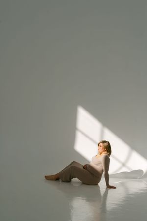 Artistic maternity photo with dramatic lighting showing a woman in beige dress reclining on white floor.