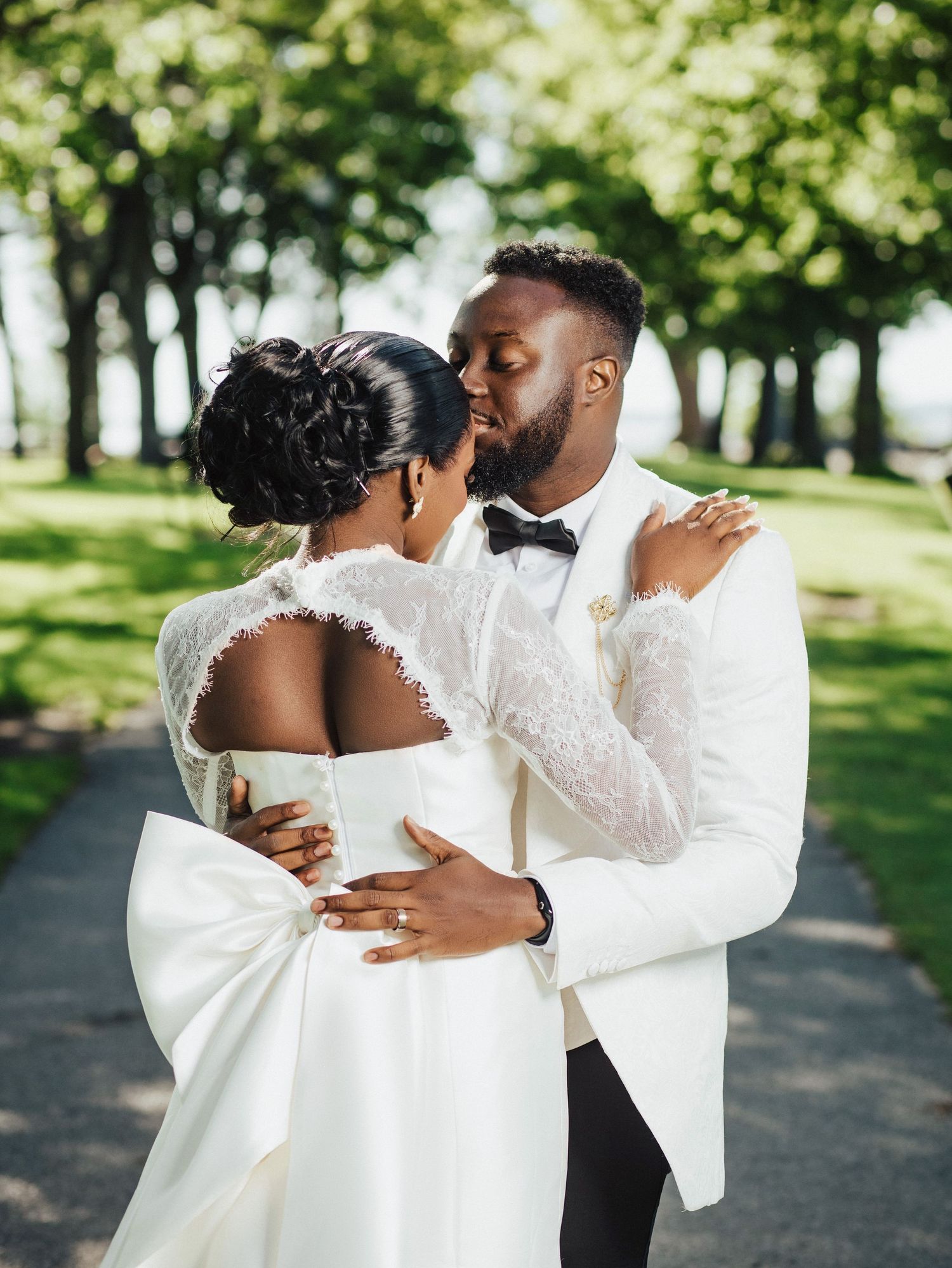 A romantic wedding moment captured in a sunlit park with newlyweds embracing in matching white attire.