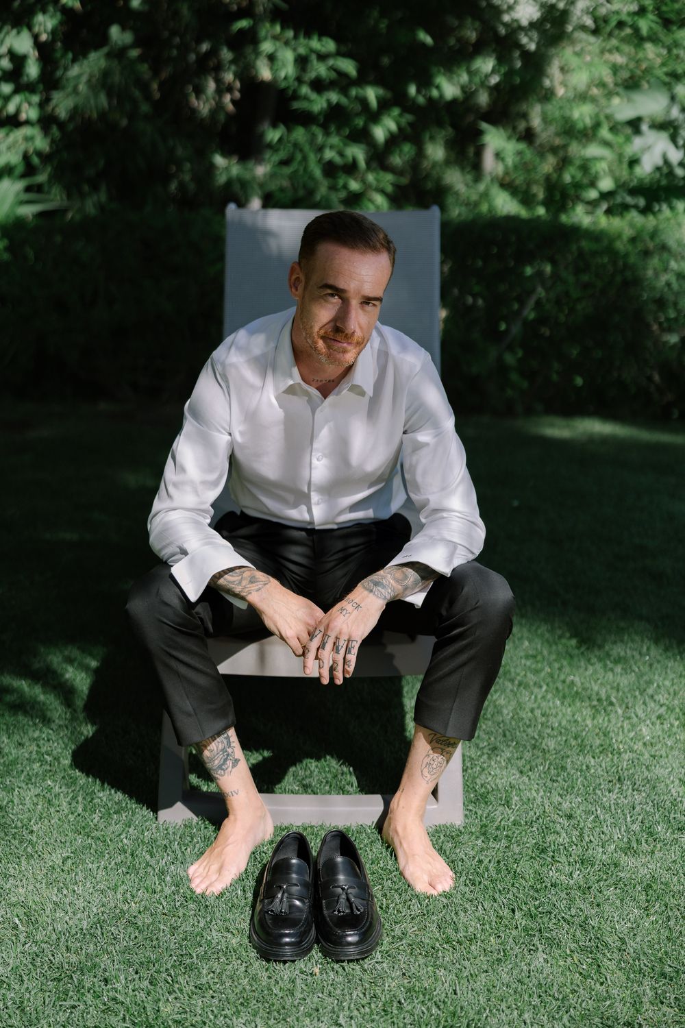 Person in white dress shirt and dark pants sits on grass next to formal black leather shoes in outdoor setting.