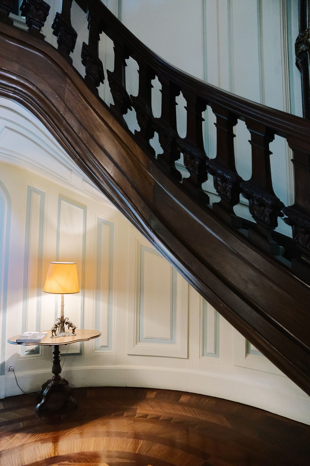 A wooden bannister curves along a white paneled wall with a warm table lamp illuminating the elegant staircase corner.