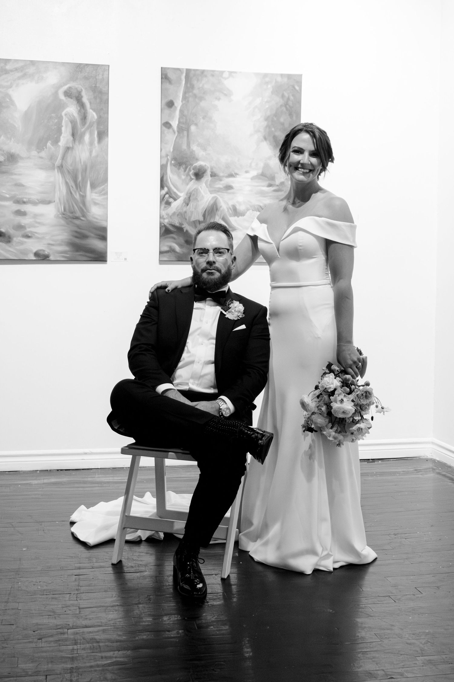 Black and white photo of a formal wedding couple posing in an art gallery with paintings visible on the wall behind them.