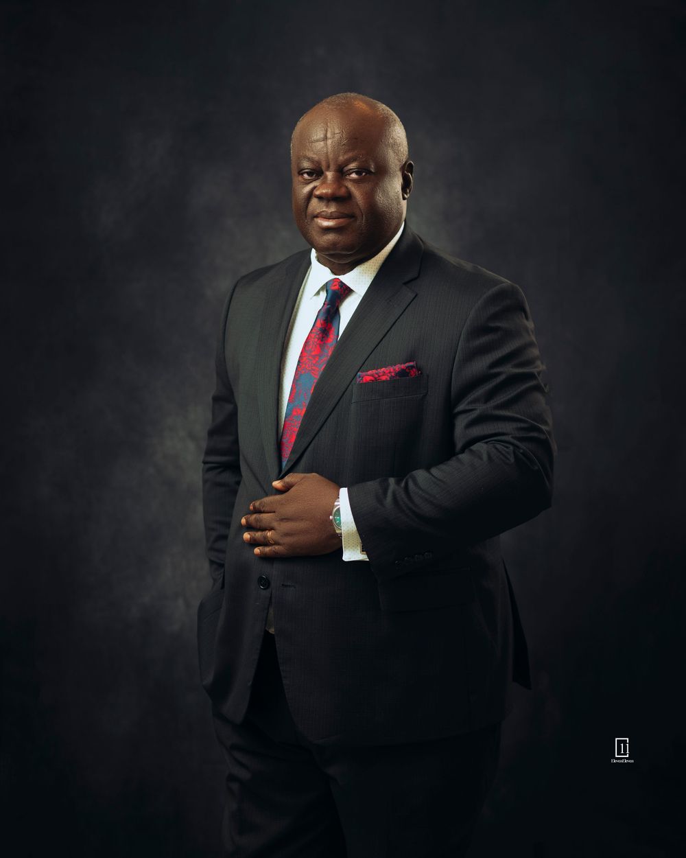 Formal business portrait of a man in dark suit and red tie against black background.