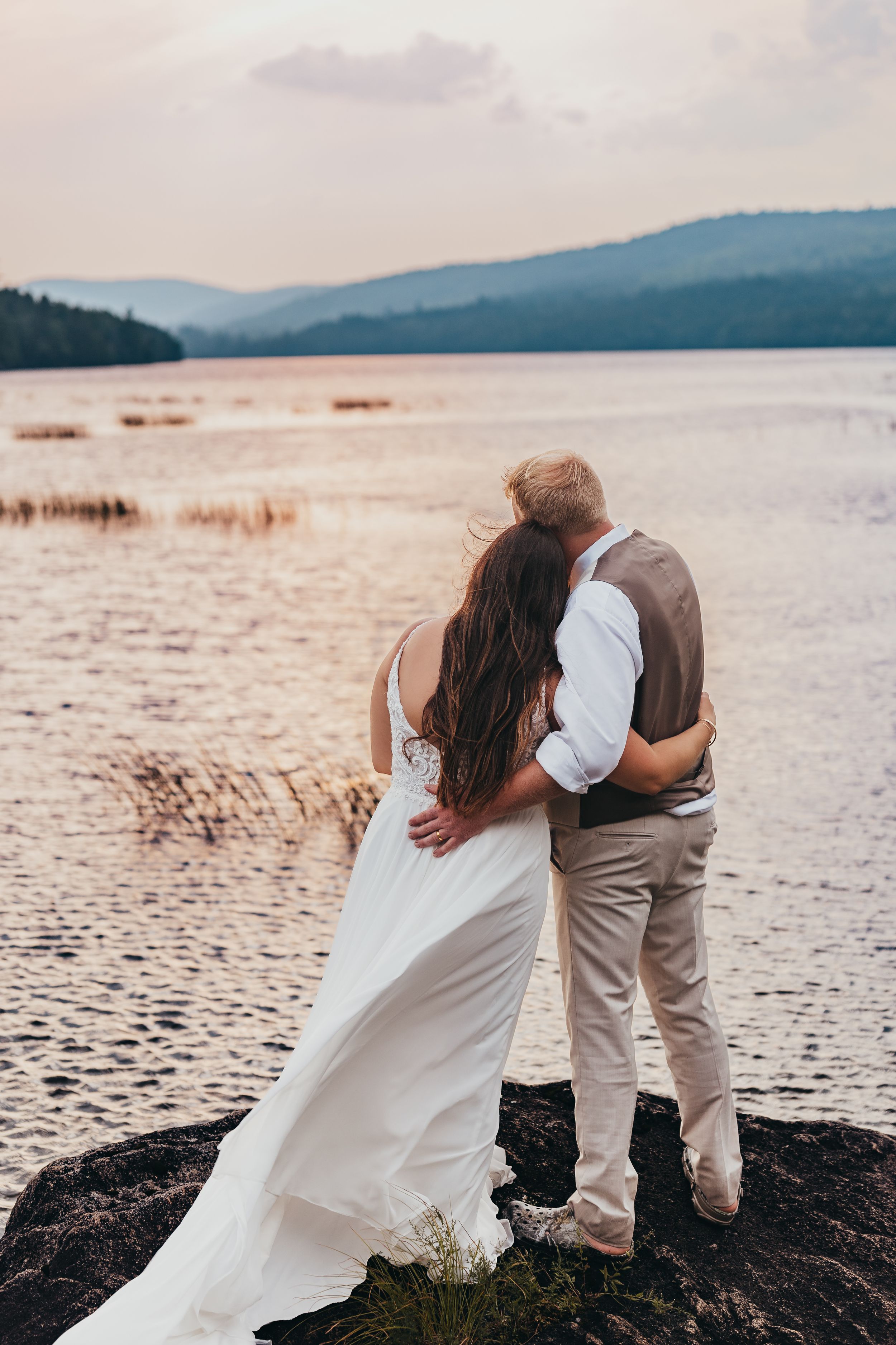 Romantic waterfront wedding photos with couple embracing on rocky shore at sunset.