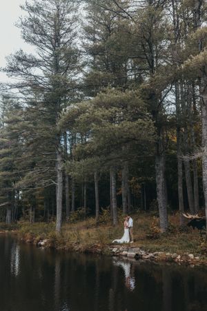 Wedding couple stands by serene lake surrounded by pine trees.