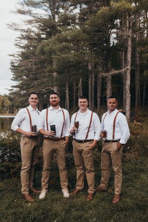 Groomsmen in matching white shirts, khaki pants and suspenders pose together in a wooded outdoor setting.