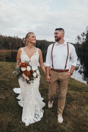 A couple takes wedding photos by a lake with the bride in lace dress and groom in casual attire.