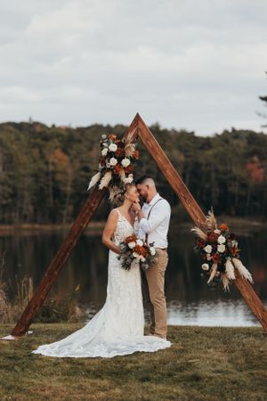 Wedding couple poses in front of a wooden triangle arch with floral arrangements by a lake.