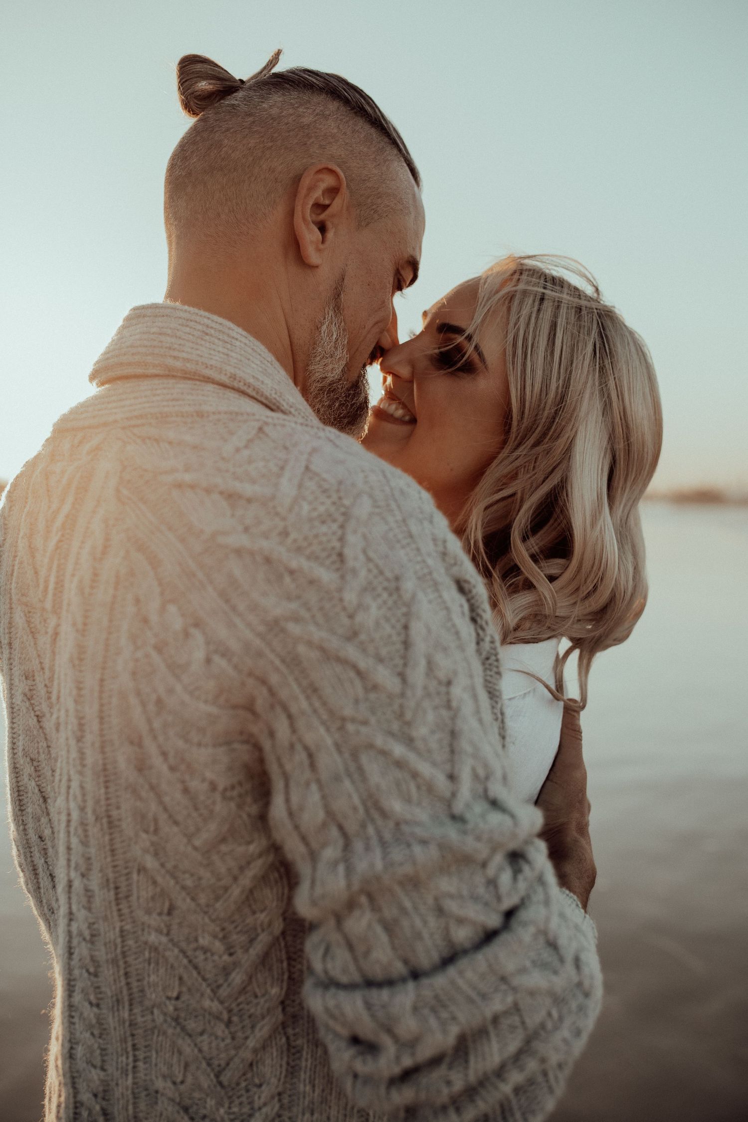 A romantic couple shares an intimate moment at sunset while wearing cozy sweaters on the beach.