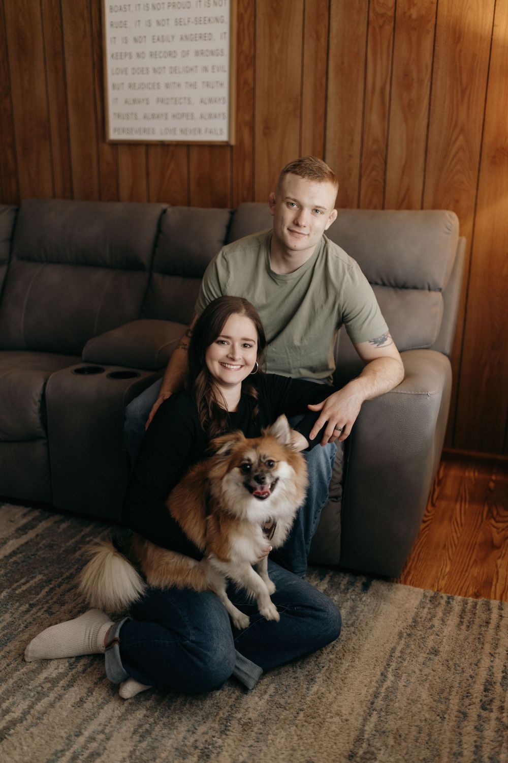 A couple sits on a gray couch with their fluffy Pomeranian dog in a wood-paneled room.
