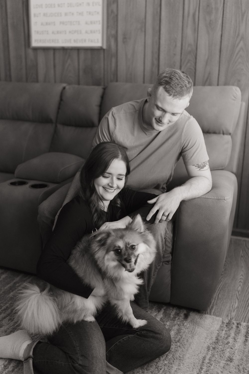 A black and white portrait of a couple relaxing with their Pomeranian on a gray sofa.