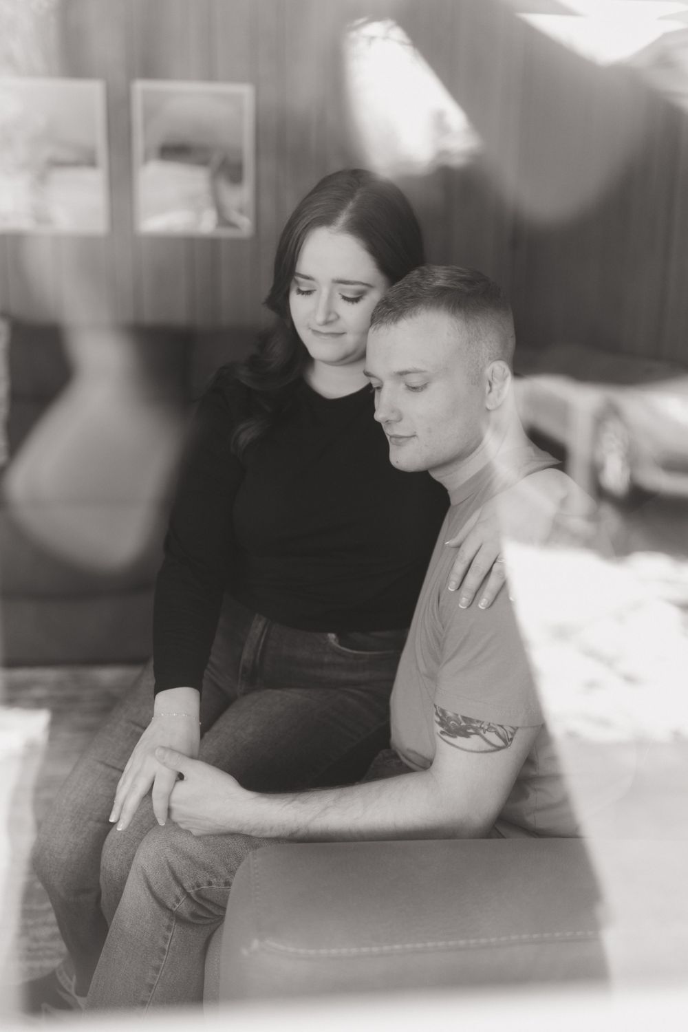 Intimate black and white photo of couple sitting close together on couch in atmospheric lighting.