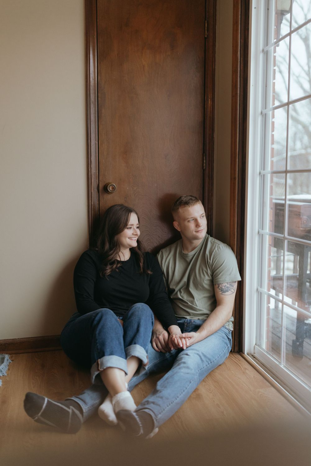 Couple relaxing by large window in casual attire with natural lighting.