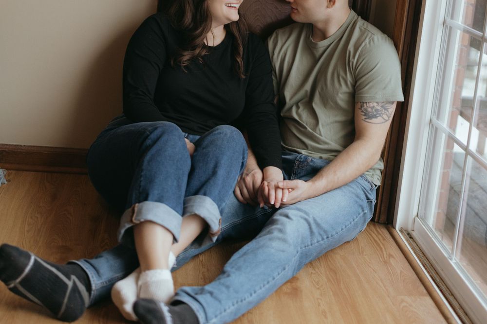A couple sits intimately together on a hardwood floor by a window wearing casual denim and t-shirts.