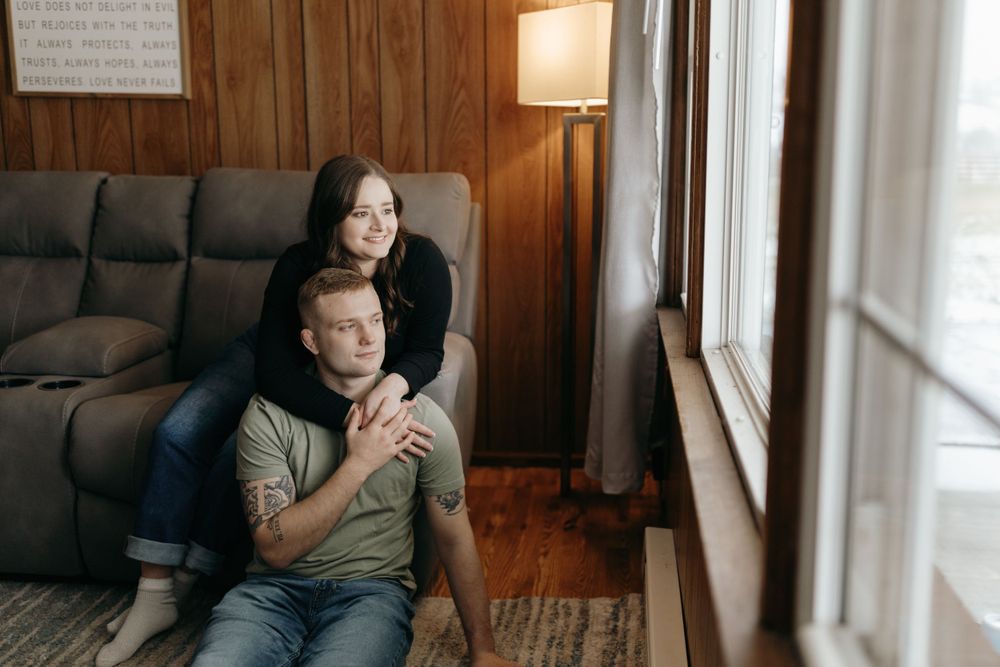 A couple poses by a window in a living room with wood paneled walls and natural light.