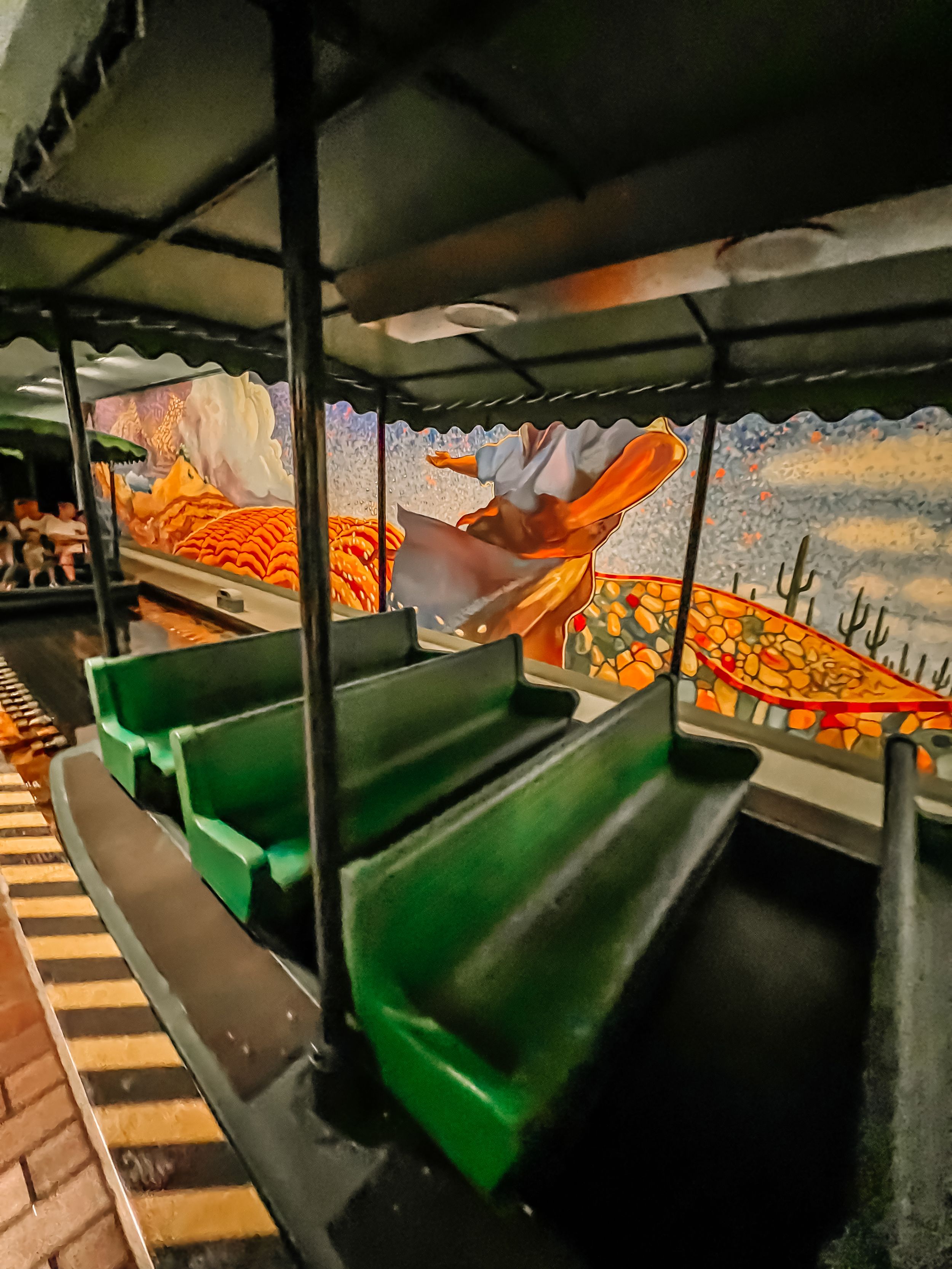 Empty seats with green cushions inside a dark amusement park ride car with colorful mural artwork on the surrounding walls.