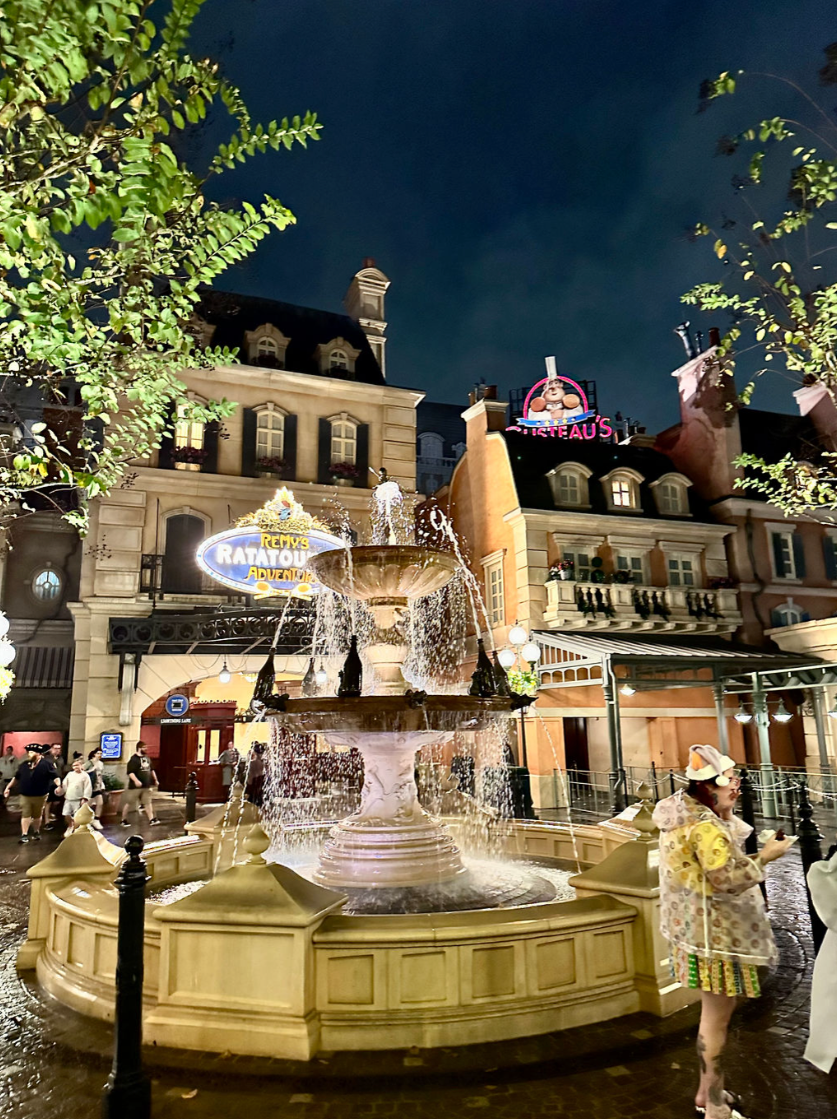 Illuminated fountain at New Orleans Square in Disneyland at night with French Quarter architecture and colorful signs.