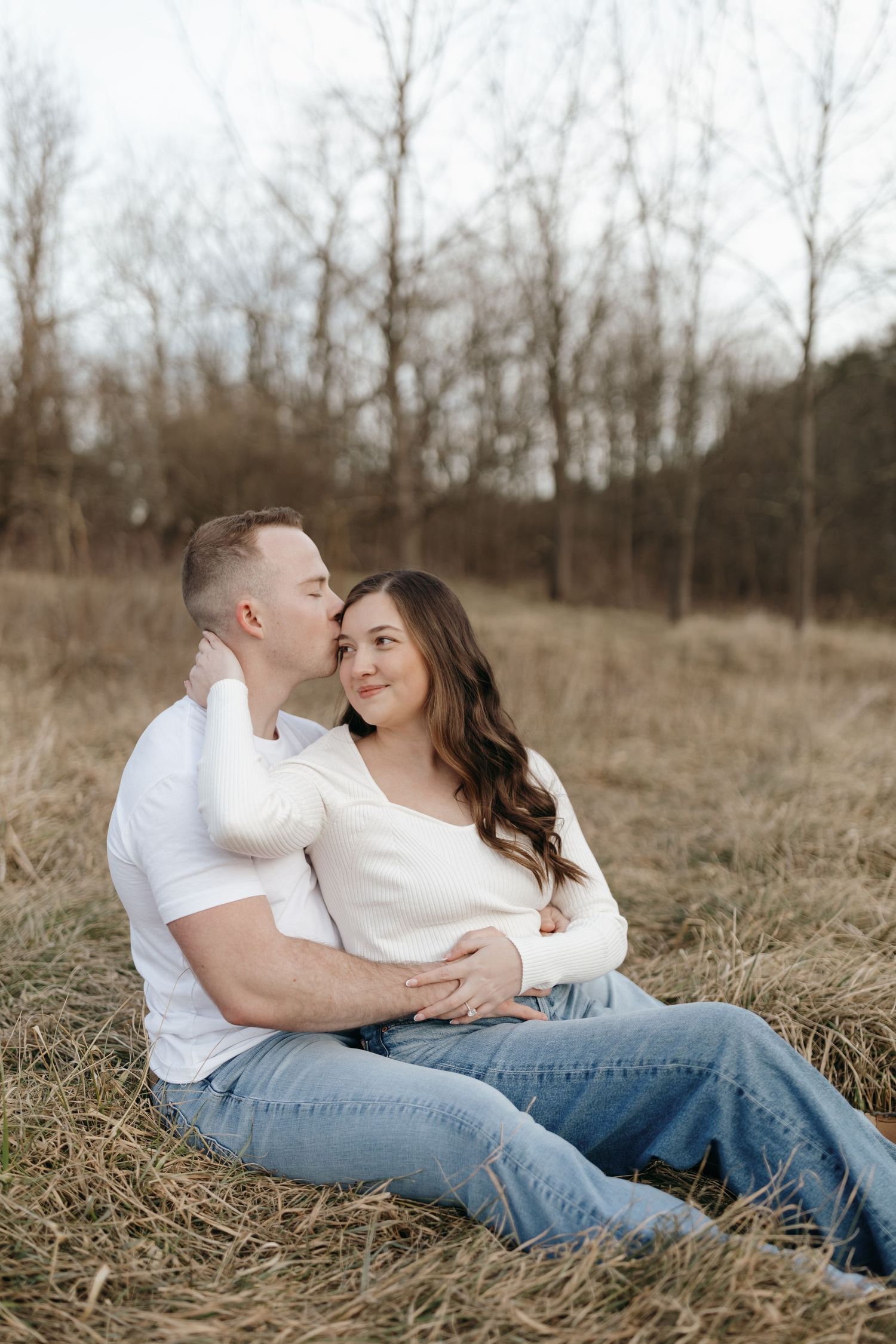 A casual couple sits intimately in a field during a winter engagement photoshoot.