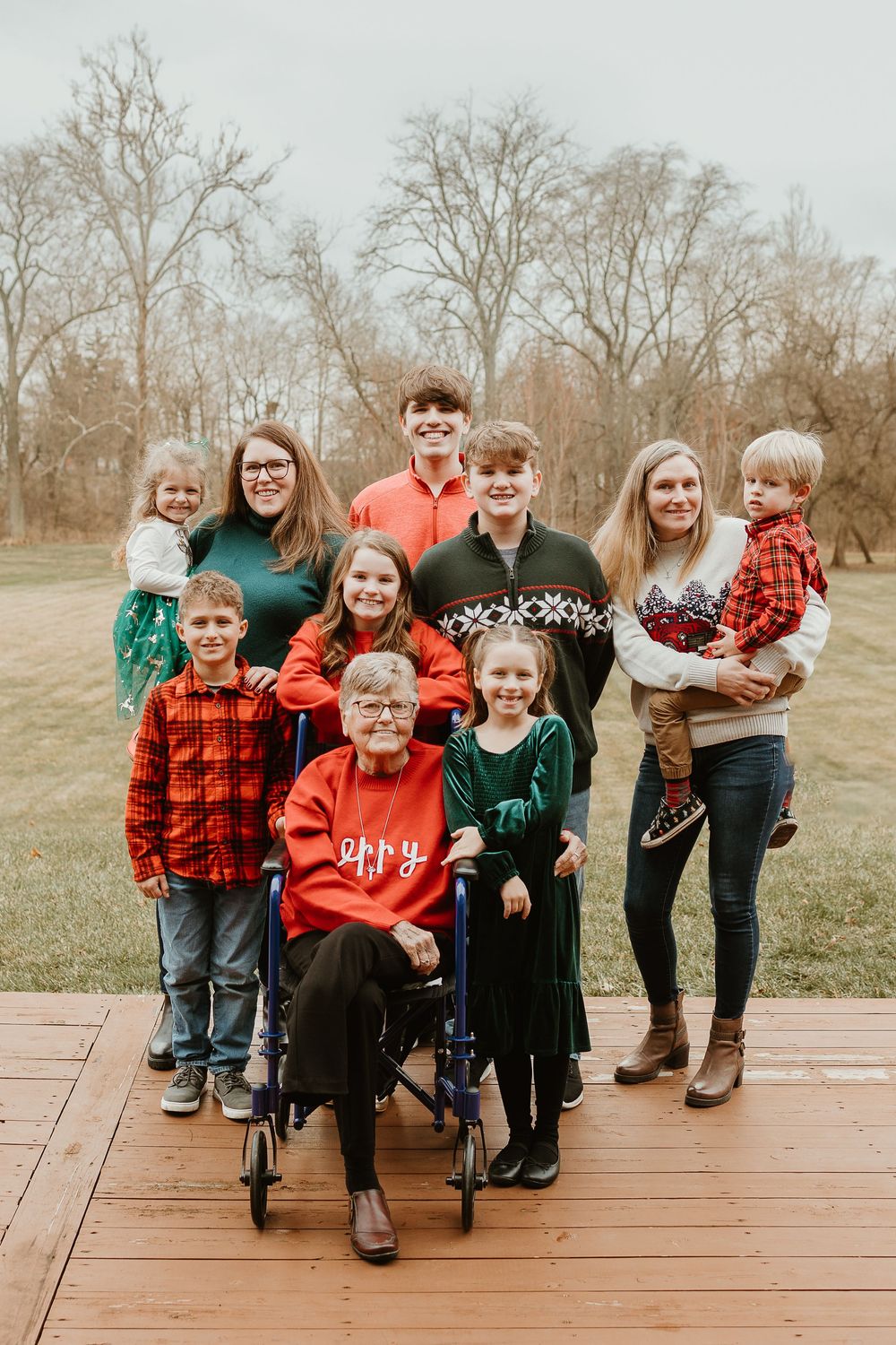 A family group poses on a wooden deck wearing coordinated red and green holiday outfits in a natural outdoor setting.