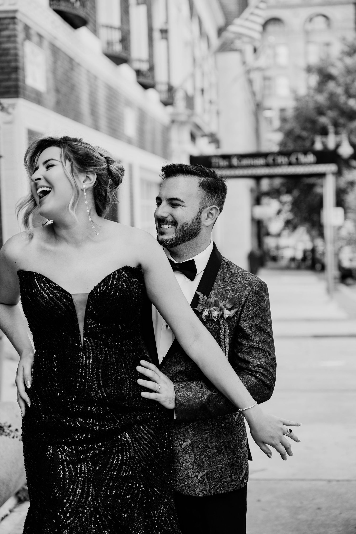 A couple laughs and embraces on a city street, with her wearing a black evening gown and him in a dark patterned suit.