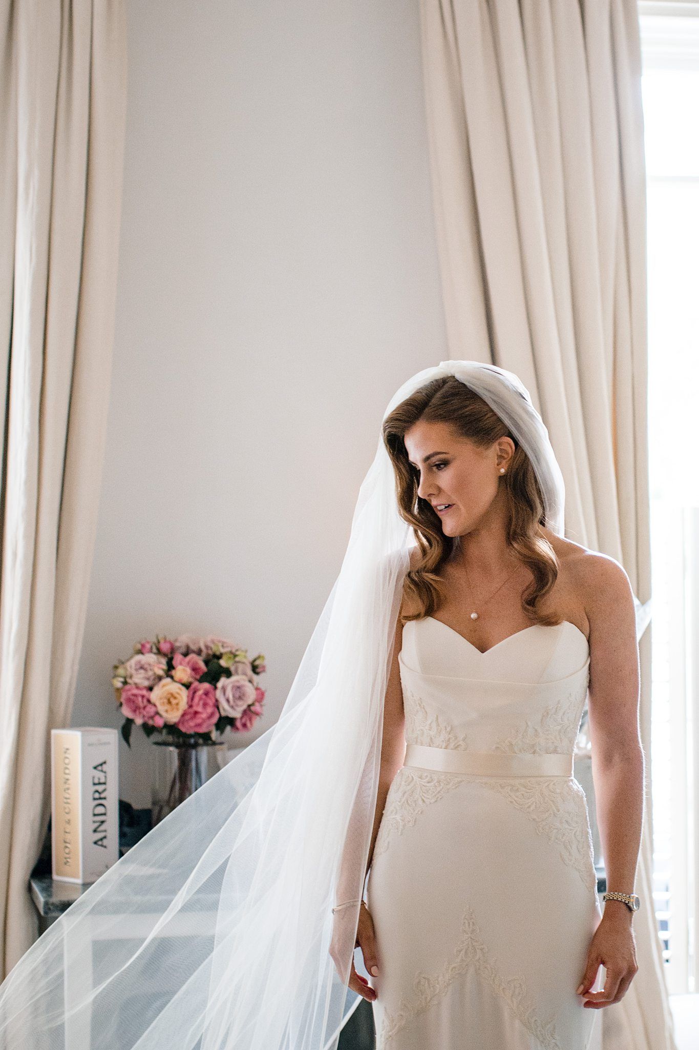 A bride in a strapless white wedding dress and long veil stands near curtained windows with pink roses nearby at The Westin Nova Scotian.