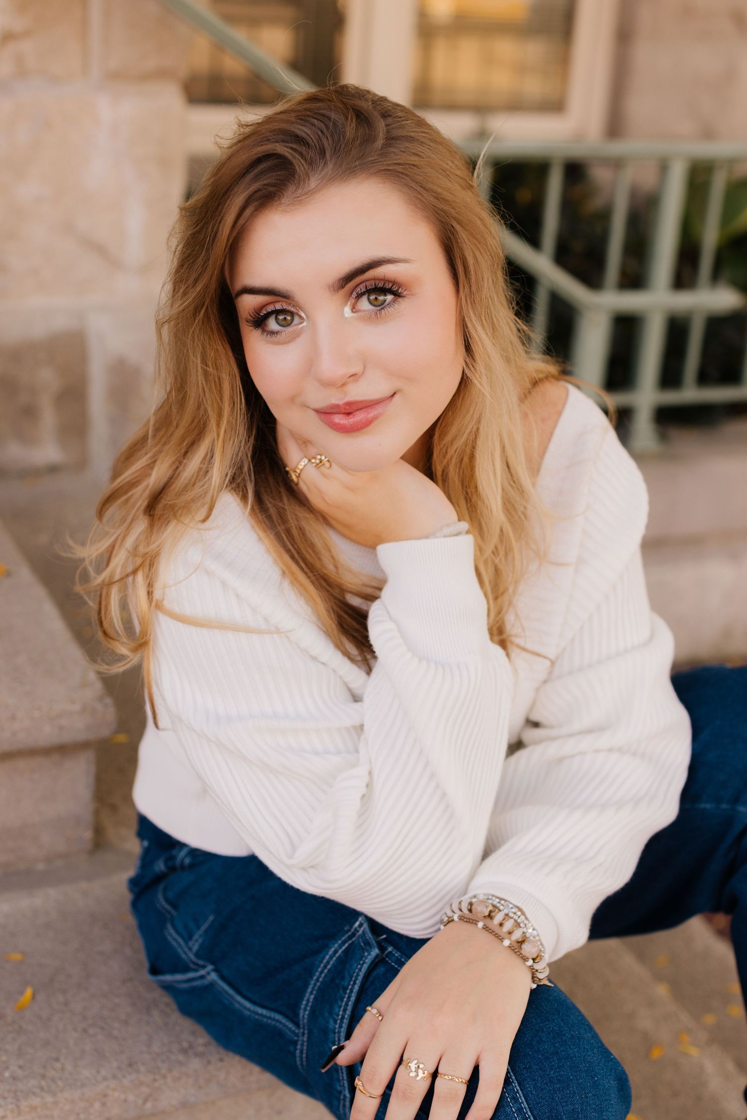 Young woman in cozy white sweater and jeans sits outdoors with hand resting on chin in warm sunlight.