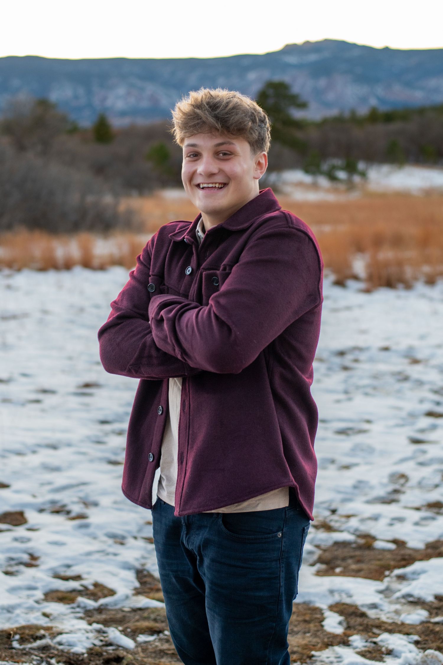 Person in a burgundy hoodie smiles with arms crossed while standing in a snowy field with mountains in background.
