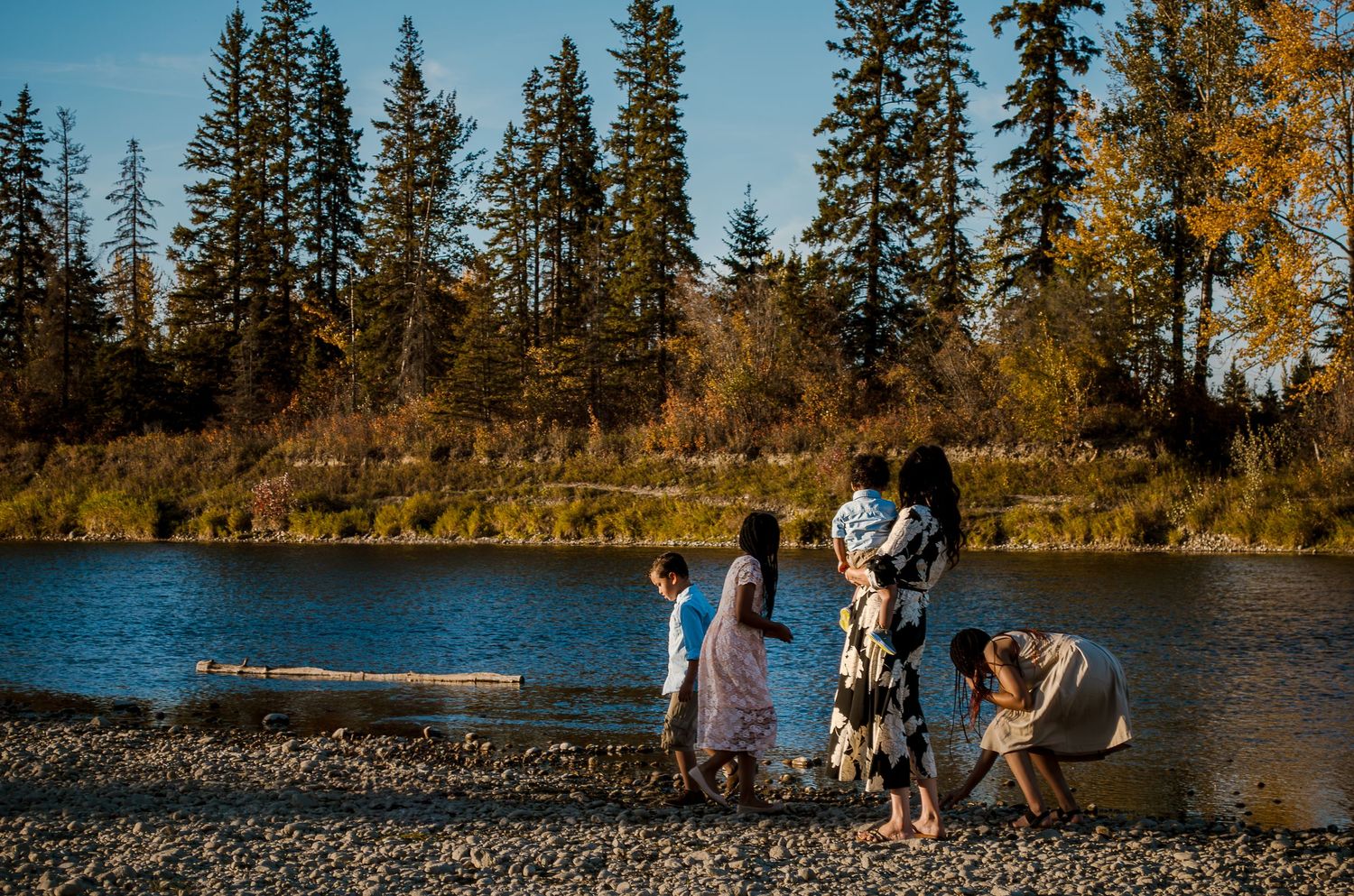 Family gathering by lakeside with evergreen trees in autumn backdrop.