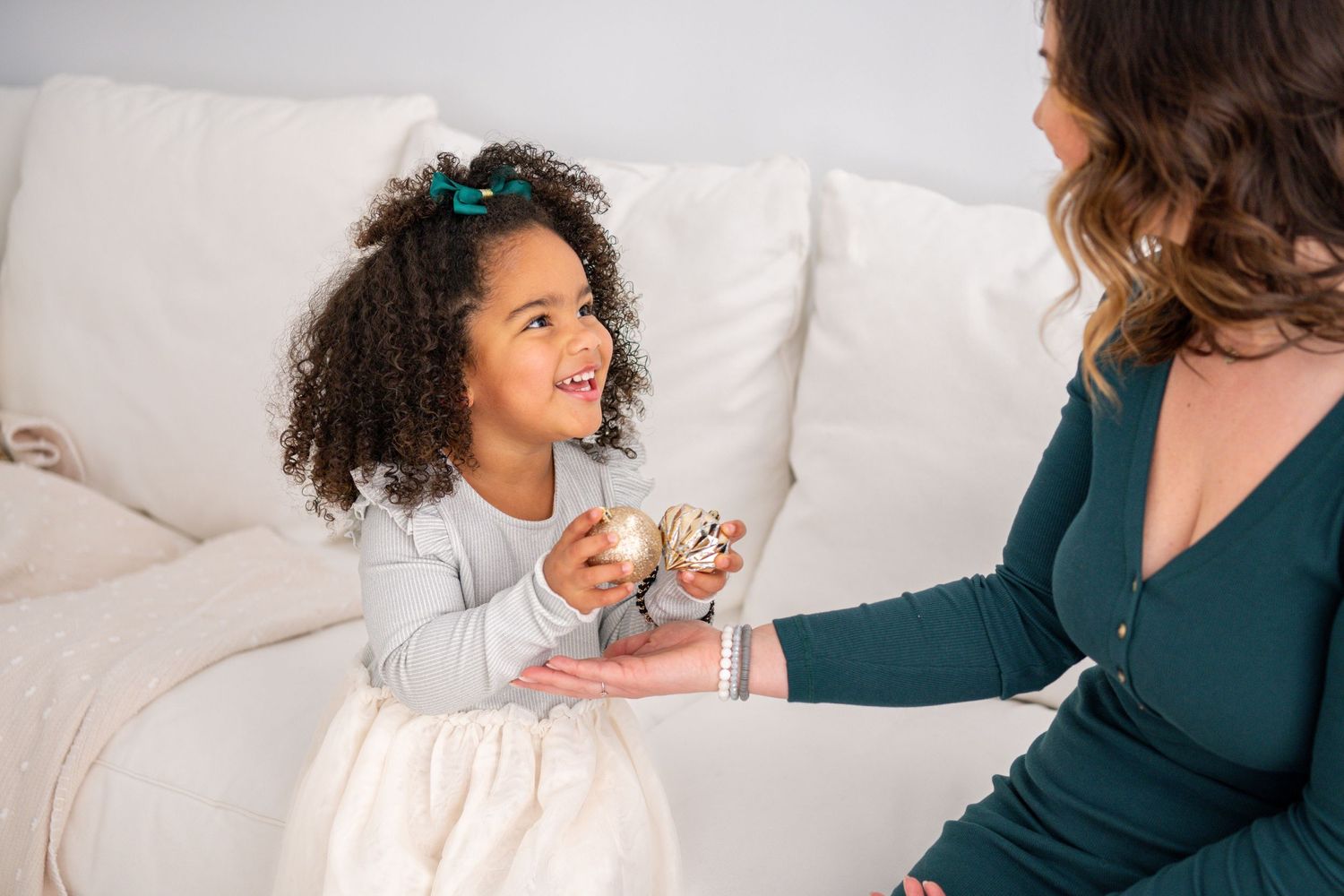 Young girl with curly hair and turquoise bow sits on white couch while interacting with woman in dark green sweater.