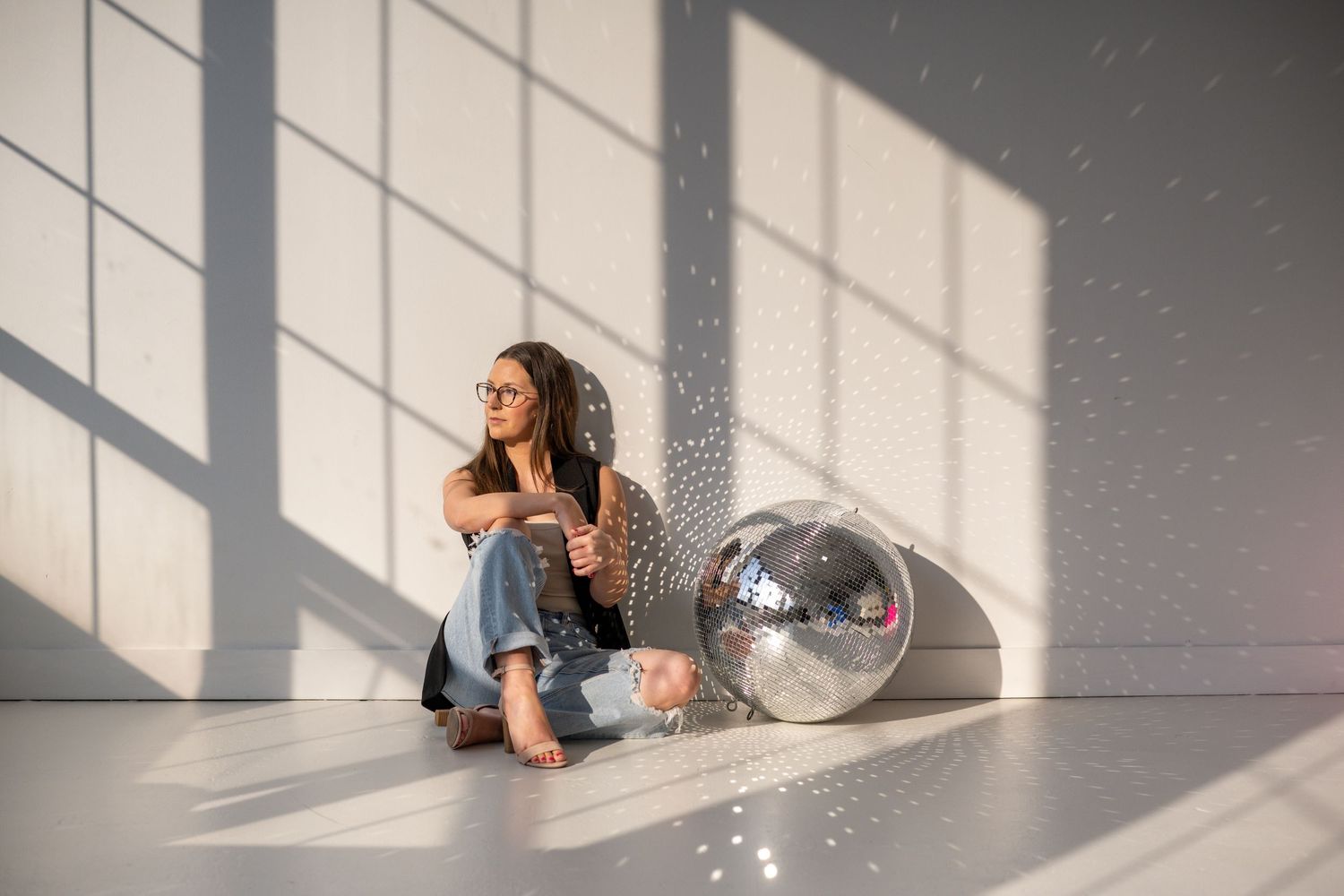 Woman sitting on floor beside disco ball while sunlight streams through windows casting dramatic shadows on white wall.