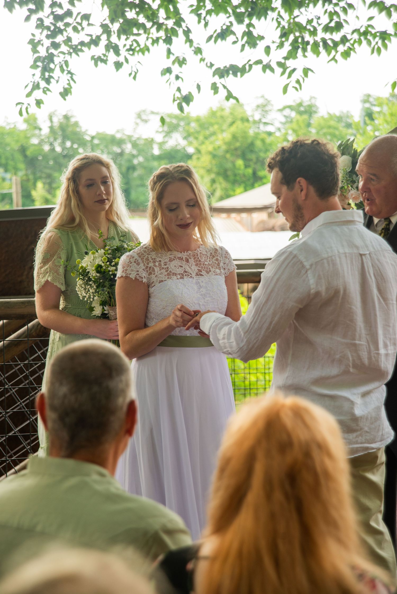 A wedding ceremony takes place outdoors with a couple exchanging rings while guests watch under green foliage.